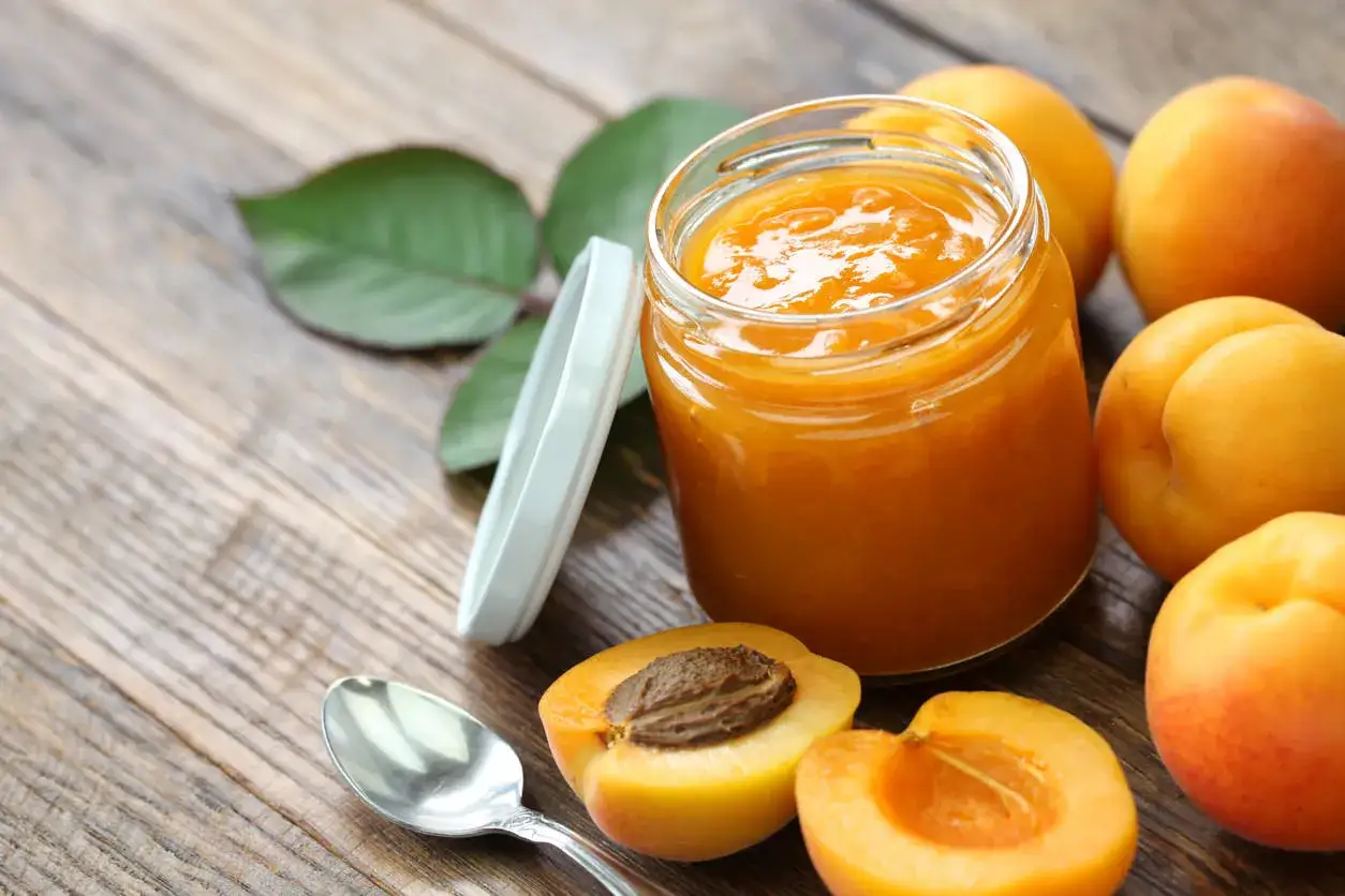 Vaso di marmellata di albicocche fatto in casa, con albicocche fresche e un cucchiaino su un tavolo di legno.