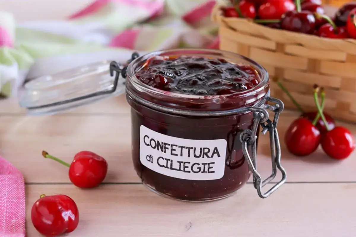 Vasetto di confettura di ciliegie fatto in casa, con ciliegie fresche sparse sul tavolo e un cestino pieno.