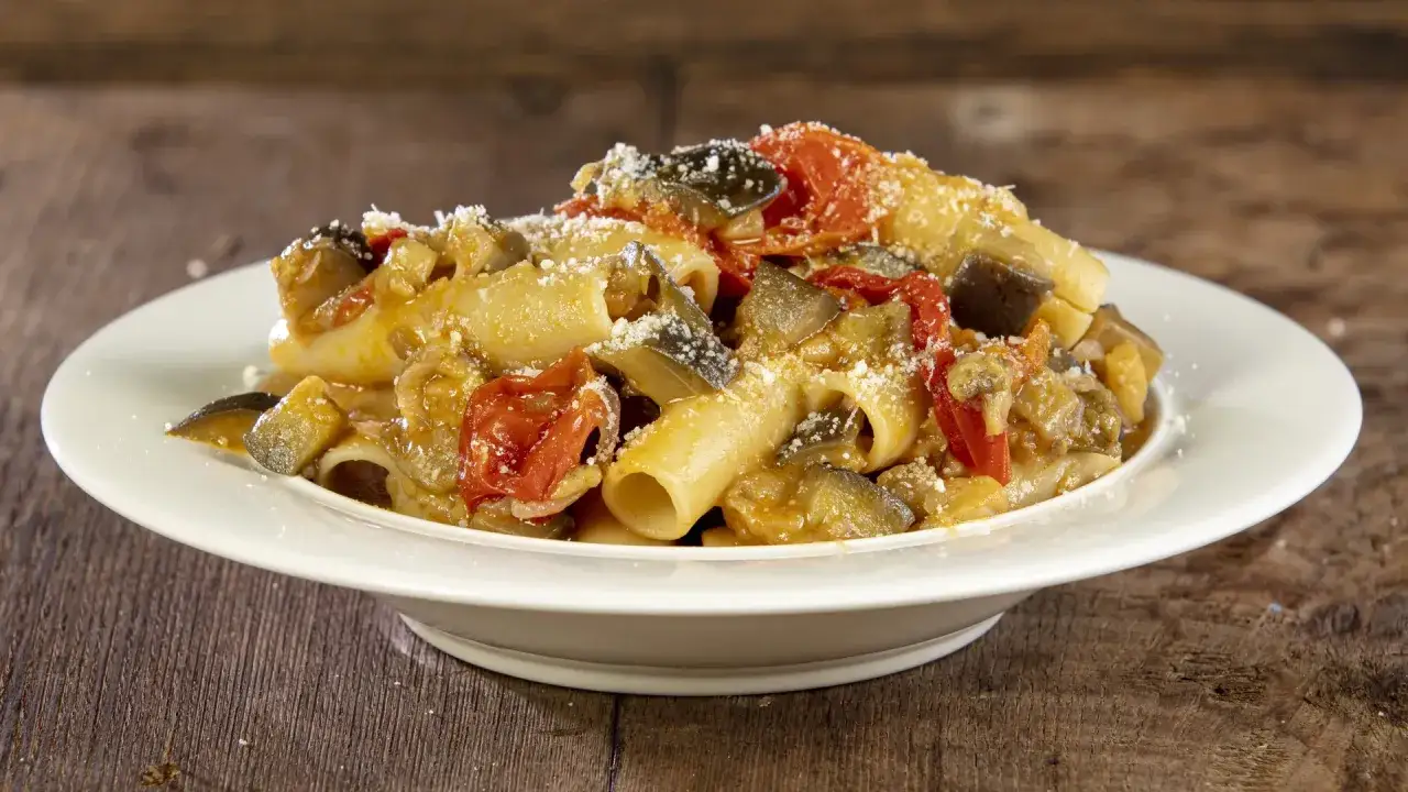 Piatto di pasta con melanzane, pomodorini e formaggio grattugiato, servito su un piatto bianco.