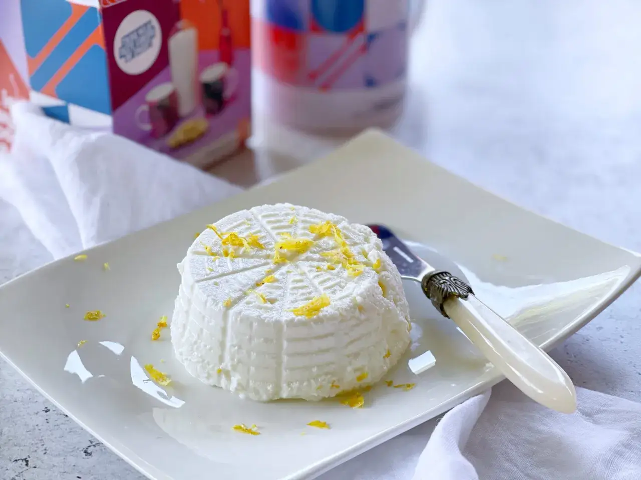 Ricotta fatta in casa, fresca e profumata, guarnita con scorza di limone su un piatto bianco.