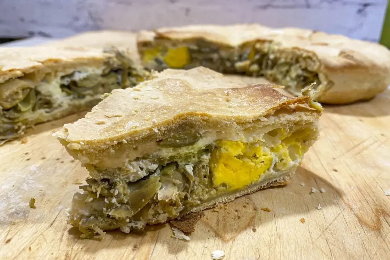 Fetta di torta pasqualina ai carciofi, con uova intere e ricotta, su tagliere di legno.