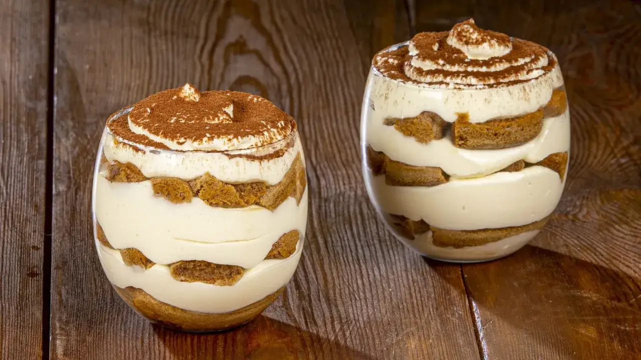 Due bicchieri di tiramisù senza uova, stratificati con crema e savoiardi, spolverati di cacao su un tavolo di legno.