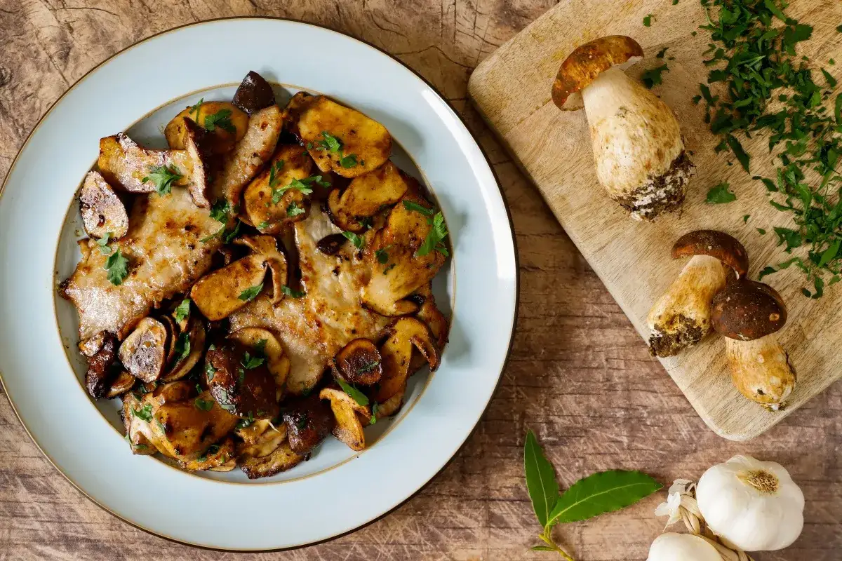 Scaloppine ai funghi su un piatto azzurro, con funghi porcini freschi e prezzemolo tritato su un tagliere.
