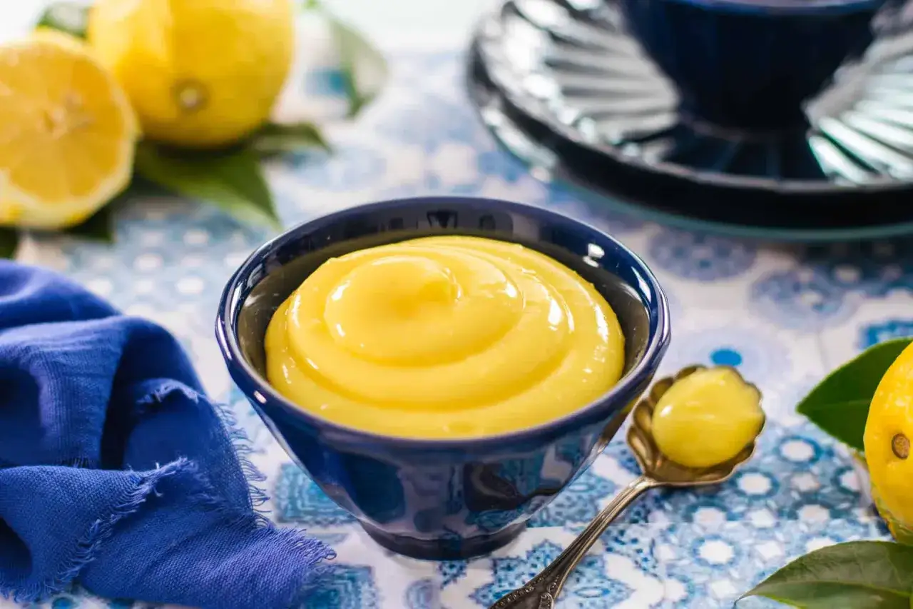 Crema al limone senza uova, liscia e vellutata, in una ciotola blu. Limoni freschi e un tovagliolo blu completano la scena.