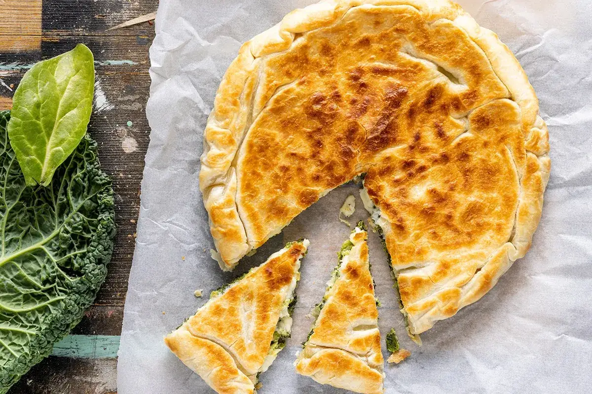 Torta salata con pasta sfoglia dorata, ripiena di verdure. Accanto, foglie di cavolo nero fresco.