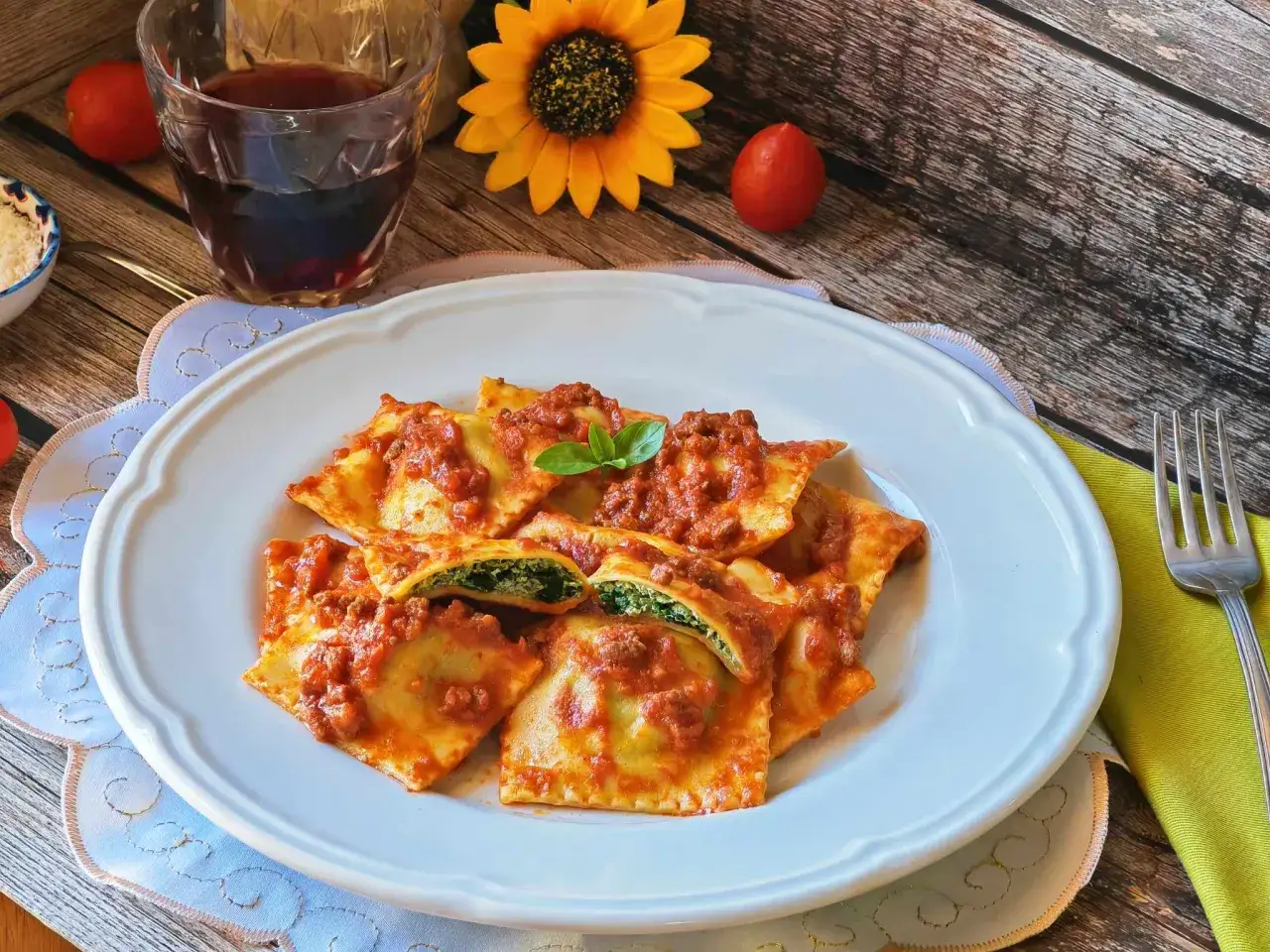 Piatto di ravioli ricotta e spinaci conditi con sugo rosso e rag&ugrave;, guarniti con basilico. Sullo sfondo, un bicchiere di vino rosso e un girasole.