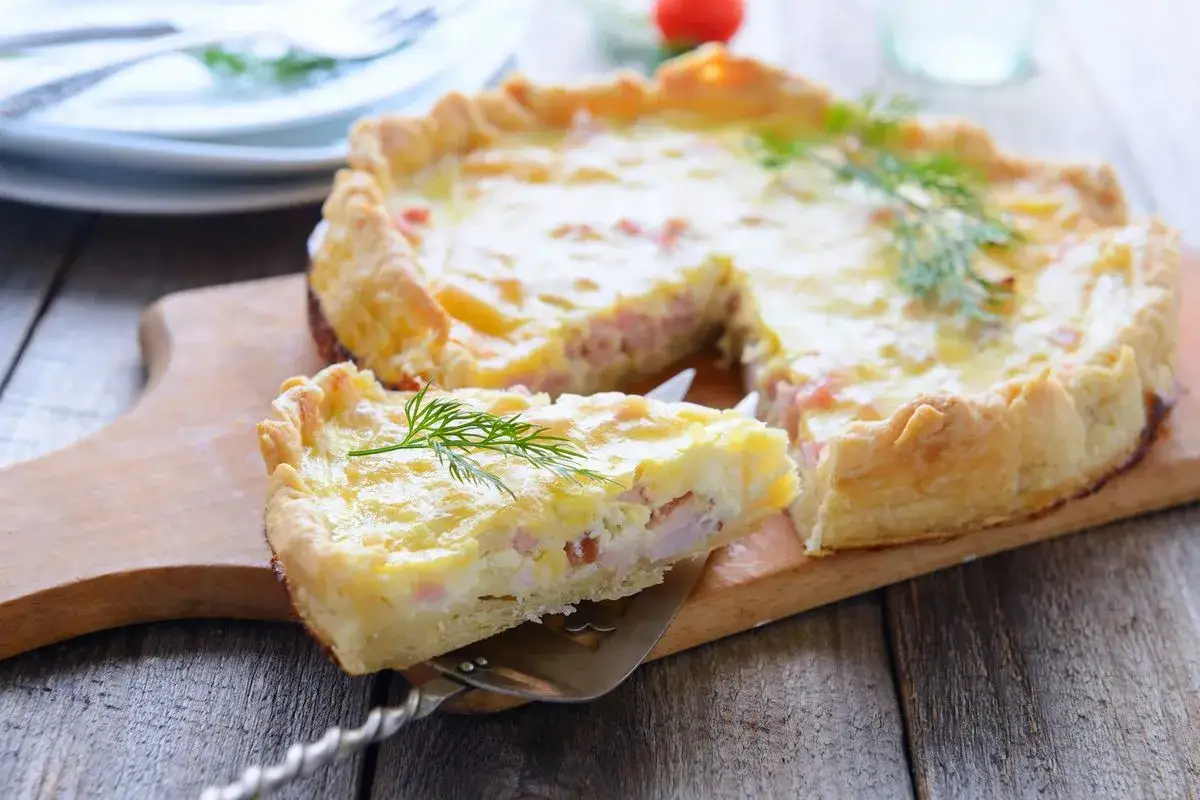 Una fetta di torta salata prosciutto e formaggio, guarnita con aneto, su un tagliere di legno.