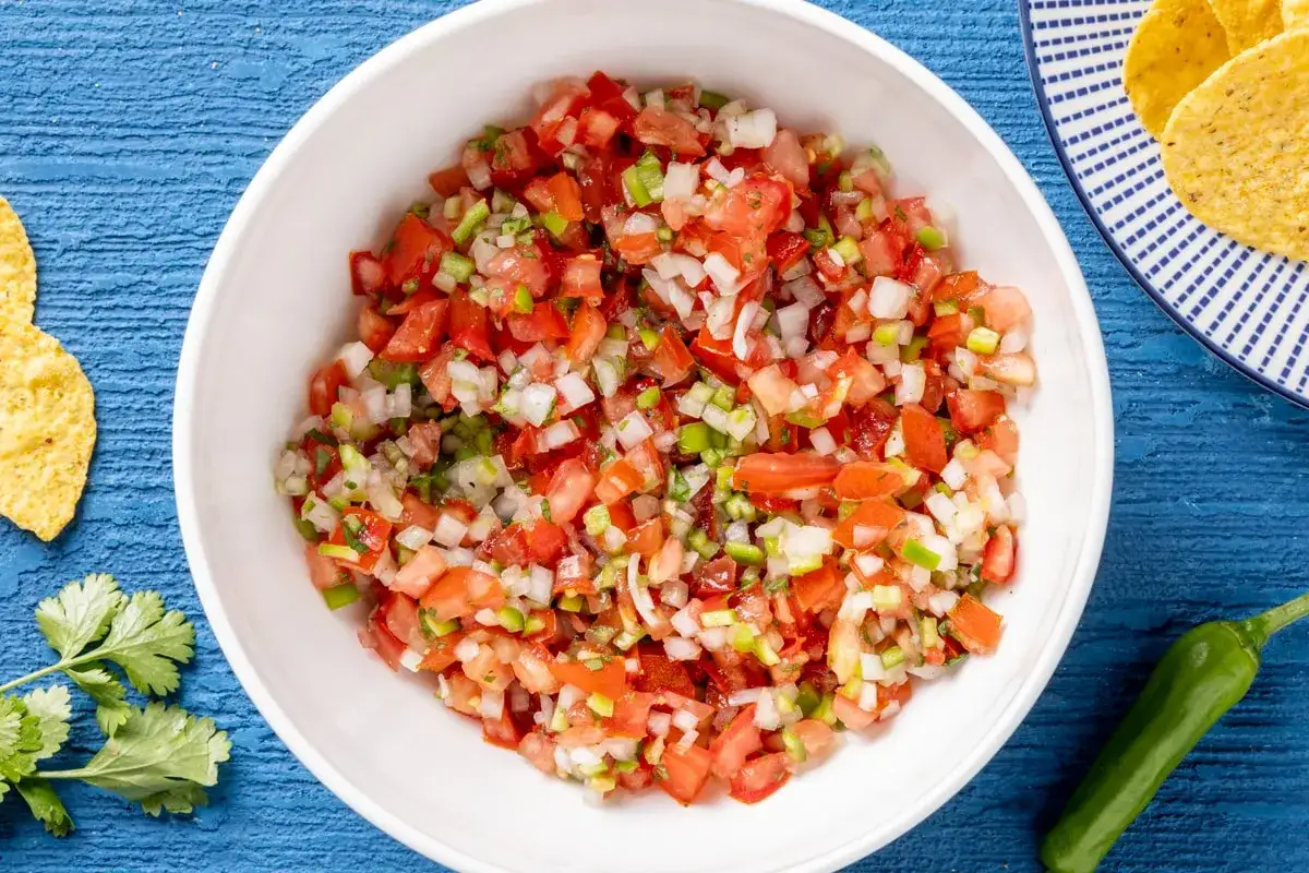 Fresca pico de gallo ricetta in una ciotola bianca, con coriandolo, peperoncino e tortillas sullo sfondo.