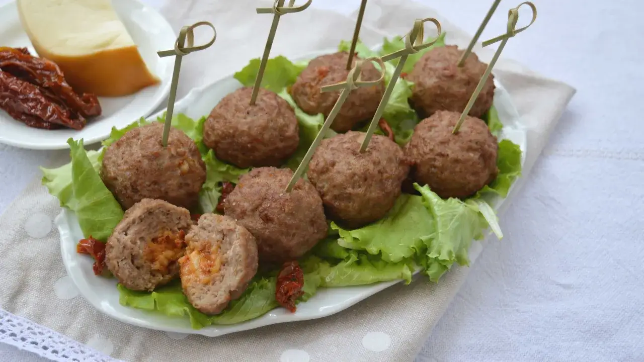 Polpette in padella servite su letto di lattuga, con pomodori secchi e formaggio sullo sfondo.