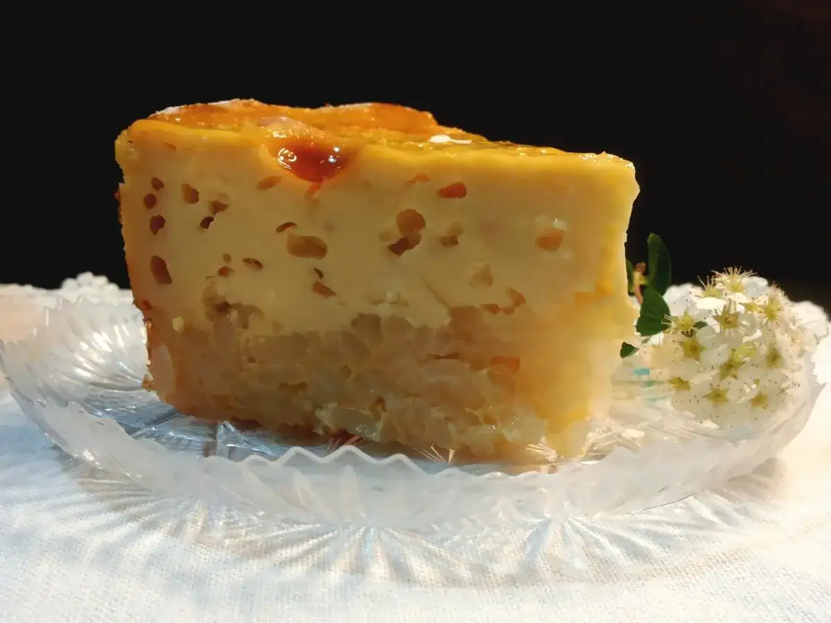 Una fetta di torta di riso carrarina, soffice e dorata, su un piattino di cristallo, con un ciuffo di fiori bianchi.
