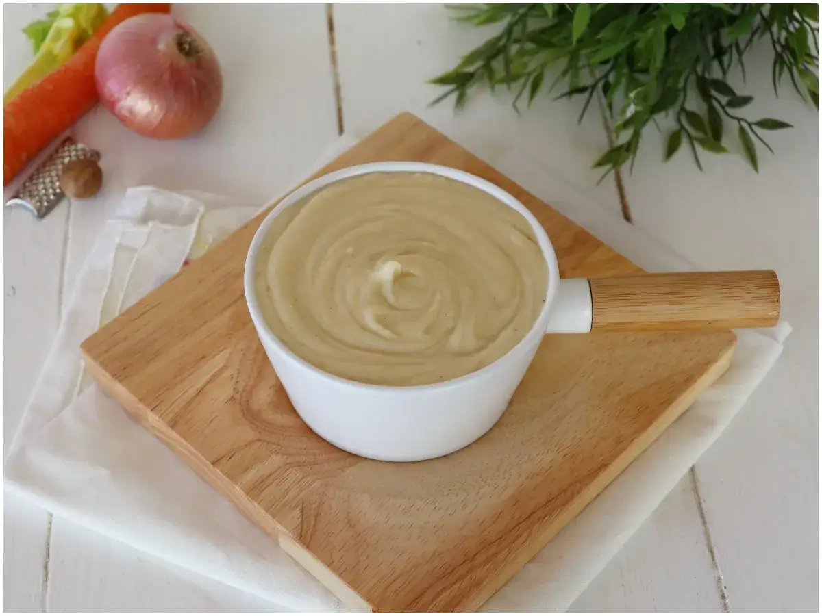 Besciamella senza latte cremosa in una ciotola bianca, con ingredienti freschi come carota, cipolla e noce moscata sullo sfondo.