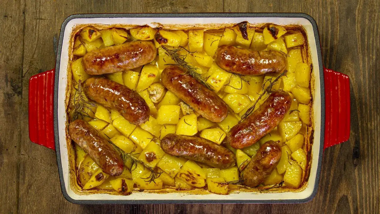 Salsiccia e patate al forno, dorate e succulente, con rosmarino e aglio, in una teglia rossa.