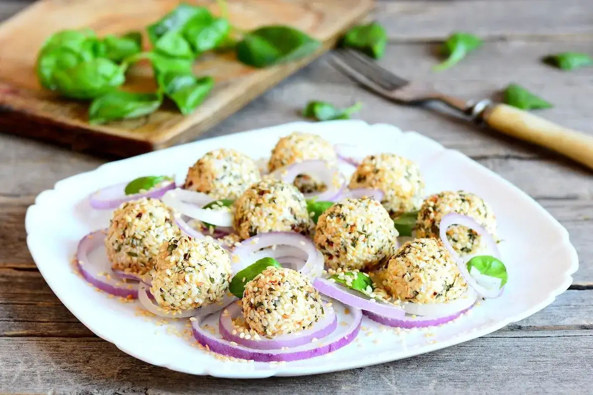 Deliziose polpette di ricotta impanate con semi di sesamo e erbe aromatiche, servite su un letto di cipolla rossa e foglie di basilico fresco.