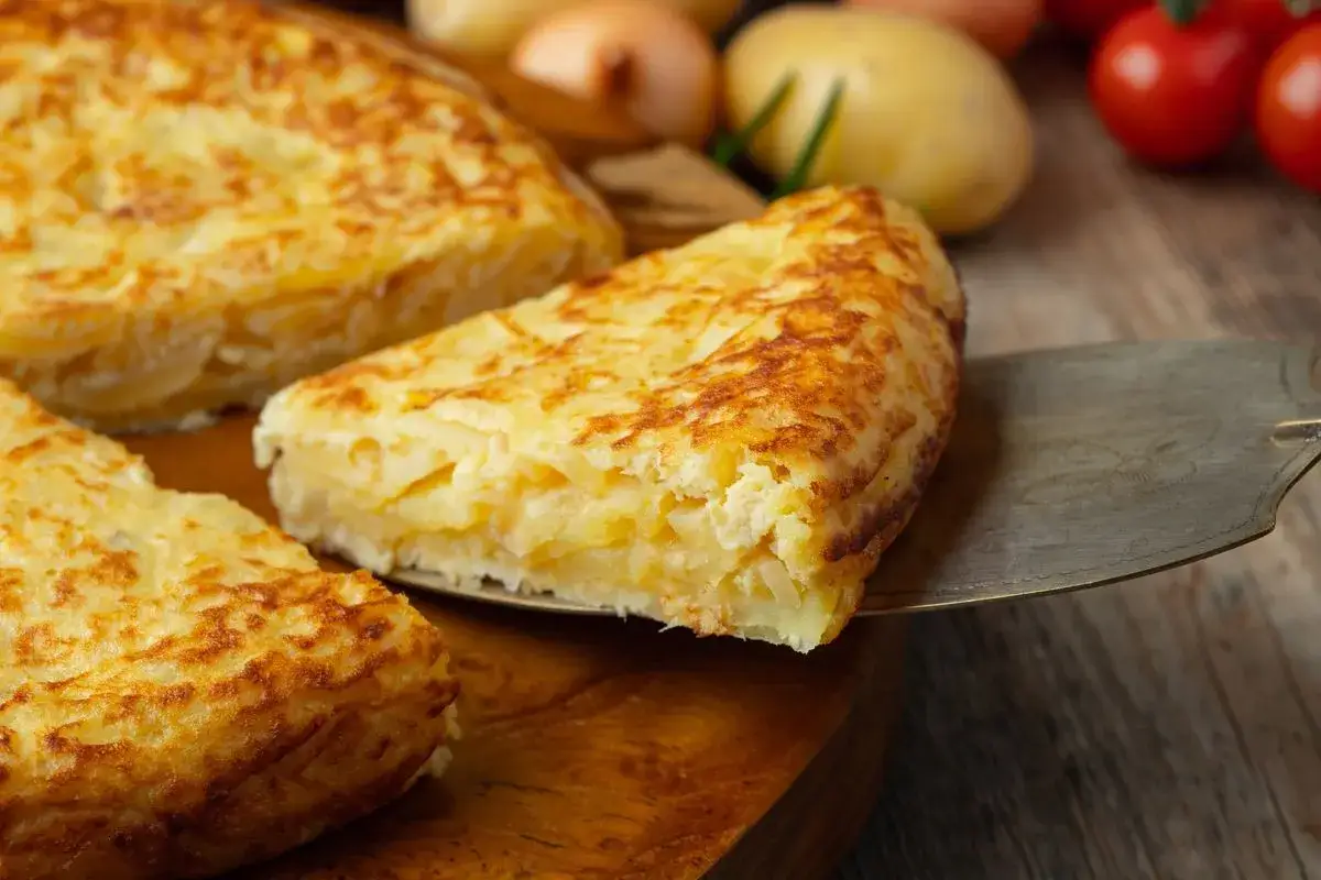 Una fetta di frittata di patate dorata viene sollevata da una tortiera su un tagliere di legno. Sullo sfondo, patate, cipolle e pomodorini.