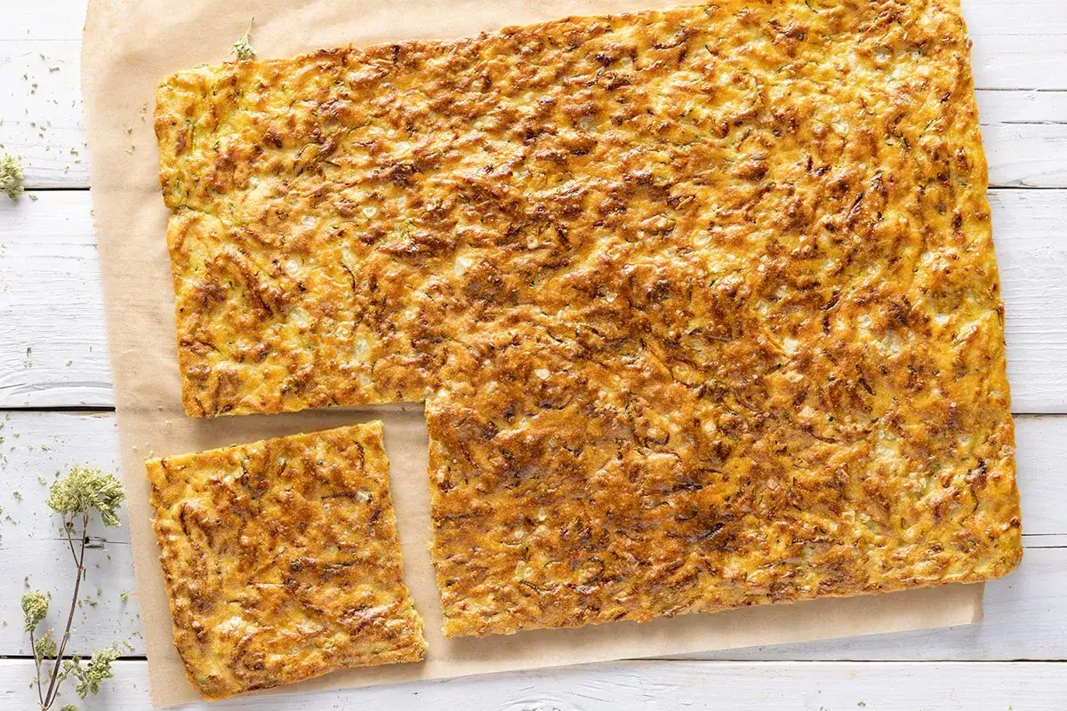 Schiacciata di zucchine dorata e fragrante, tagliata a pezzi su carta da forno, con erbe aromatiche sparse.
