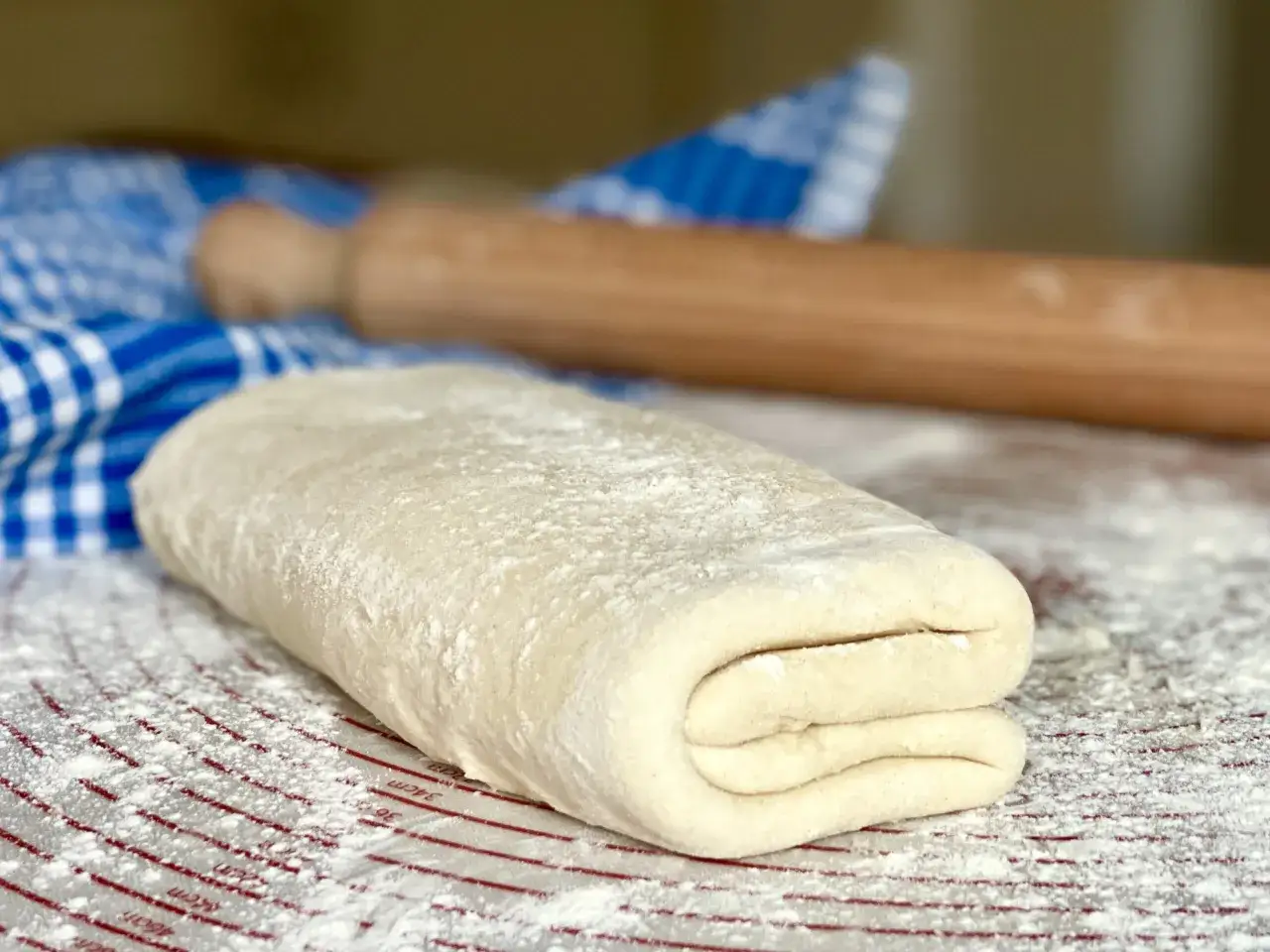 Impasto per pasta fatta in casa, piegato e infarinato, pronto per essere steso. Sullo sfondo, un mattarello e un canovaccio a quadri blu.