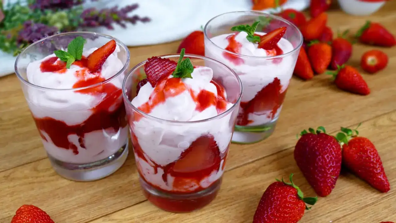 Tre bicchieri di deliziosa mousse di fragole, guarniti con fragole fresche e menta, su un tavolo di legno con fragole sparse.