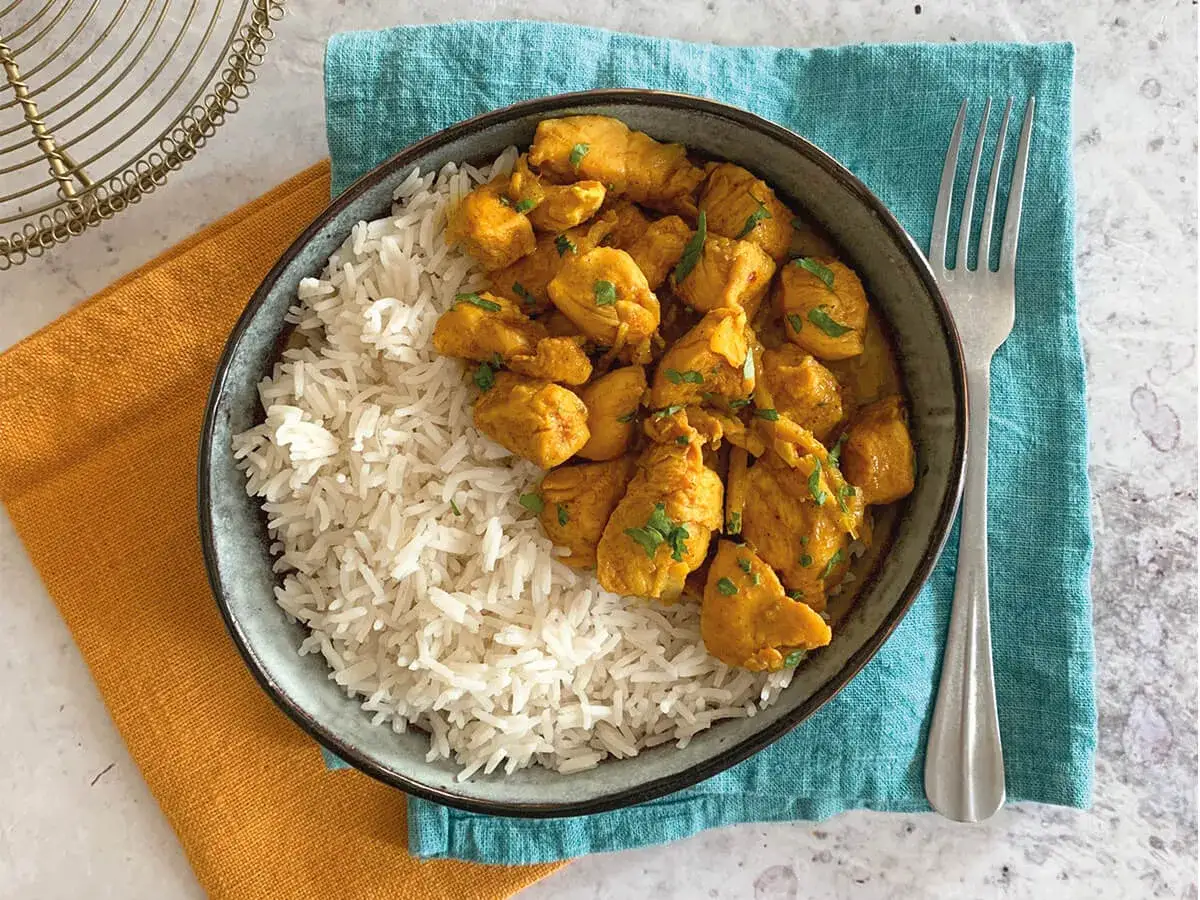Un invitante piatto di pollo al curry con riso basmati, guarnito con prezzemolo fresco. Accanto, una forchetta su un tovagliolo turchese.
