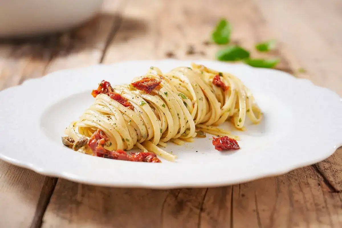 Un piatto di linguine condite con pomodori secchi, capperi e prezzemolo, su un piatto bianco bordato.