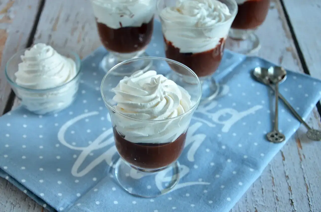 Delizioso budino fatto in casa al cioccolato, guarnito con panna montata fresca, servito in coppette di vetro su un tovagliolo a pois.