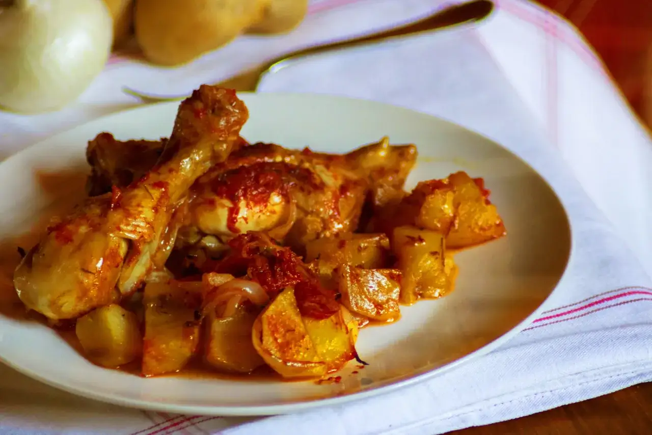 Piatto di pollo in umido con patate e pomodoro, servito su un tovagliolo bianco con righe rosa.