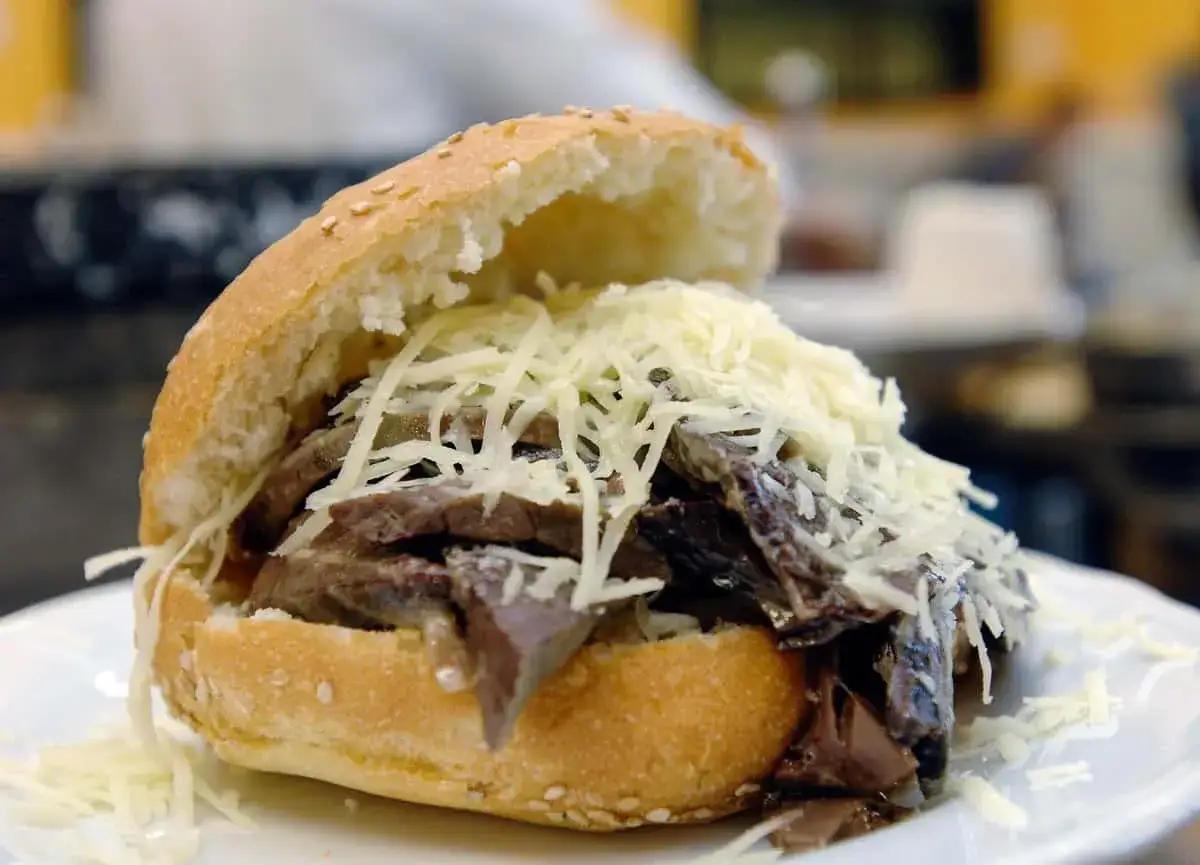 Un panino con la milza, ricco di carne scura e sfilacciata, coperto da abbondante formaggio grattugiato.