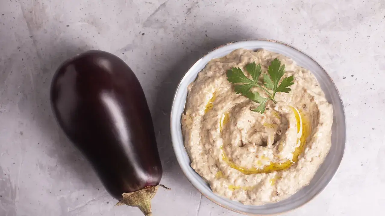 Una melanzana intera accanto a una ciotola di cremosa crema di melanzane, guarnita con prezzemolo e olio d'oliva.