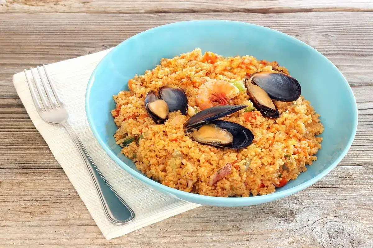Un invitante cous cous di pesce, con cozze e gamberi, servito in una ciotola azzurra su un tavolo di legno.