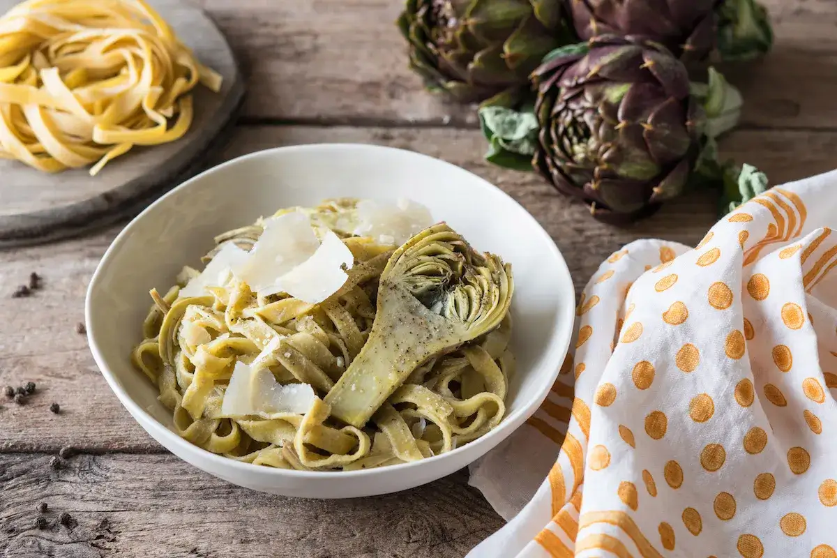 Un piatto di pasta con carciofi, guarnito con scaglie di parmigiano e pepe nero. Sullo sfondo, carciofi freschi e un rotolo di pasta cruda.