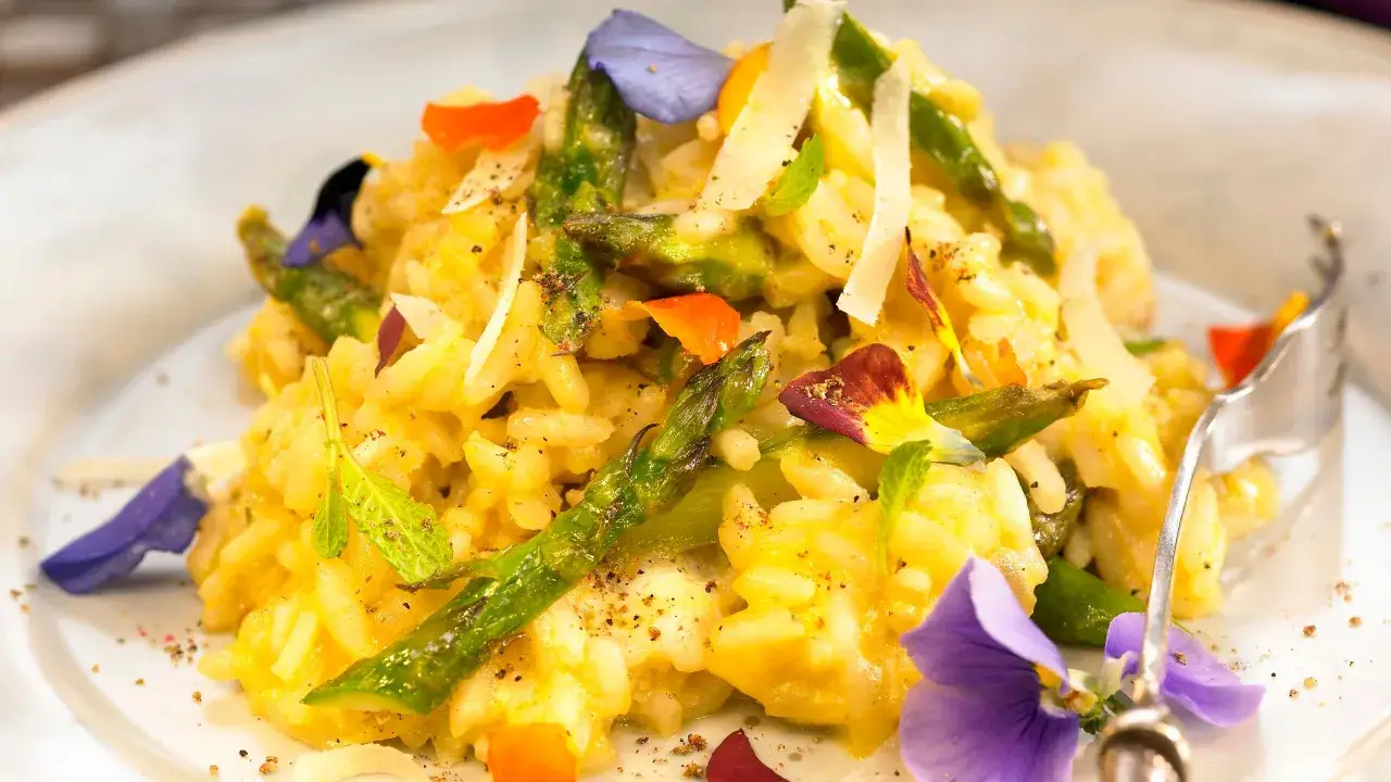 Risotto asparagi e zafferano cremoso, guarnito con asparagi freschi, scaglie di parmigiano e fiori eduli colorati.