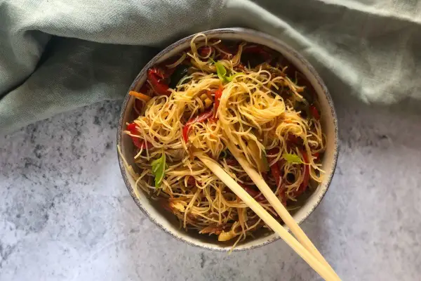 Un piatto invitante di spaghetti di riso con verdure colorate, come peperoni rossi e zucchine, guarnito con basilico fresco e servito con bacchette.