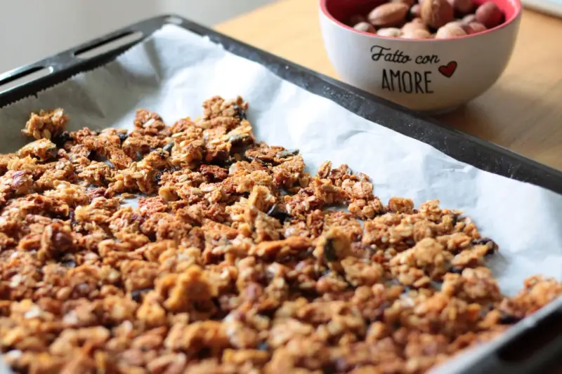 Granola fatta in casa appena sfornata su carta da forno, con una ciotola di frutta secca sullo sfondo.