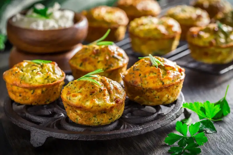 Tortini di zucchine dorati e fragranti, guarniti con rosmarino, su un piatto di ghisa. Sullo sfondo, una ciotola di formaggio fresco.